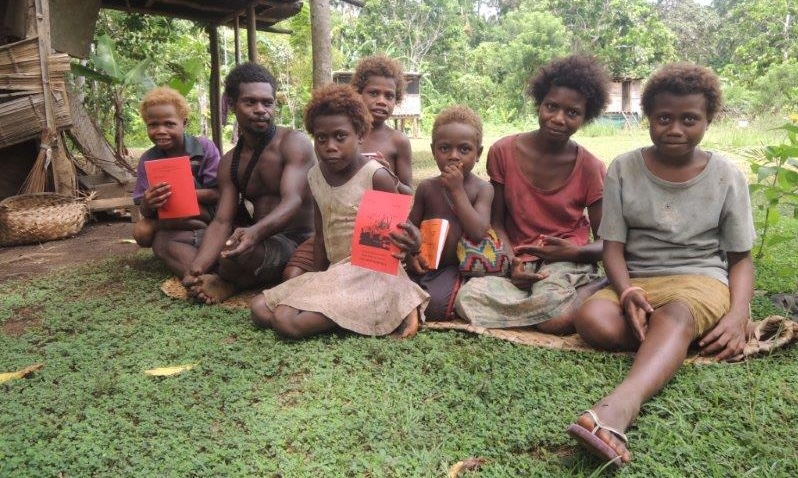Barn från Papua Nya Guinea sitter i gräset och håller upp en röd bok.